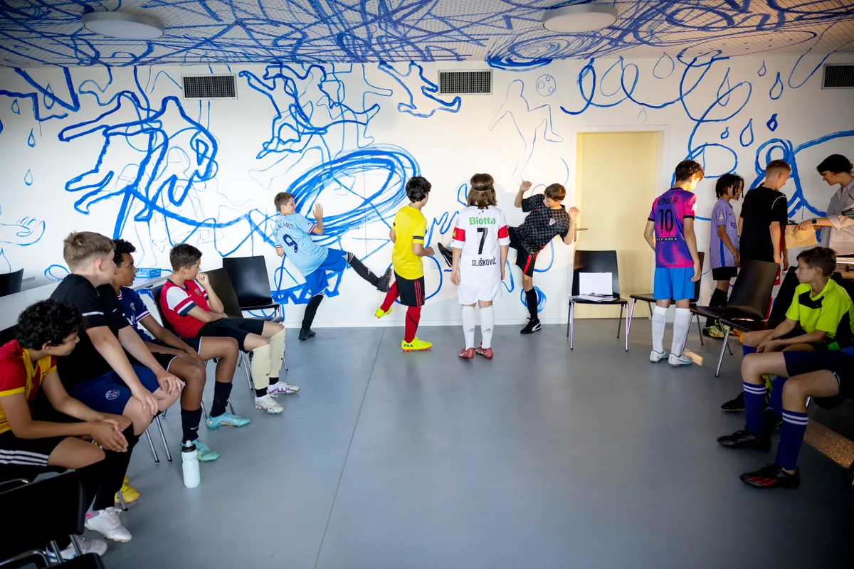 Man sieht Kunst an einer Wand in einem Raum mit Stühlen, auf denen Kinder in Fussballtrikots sitzen.